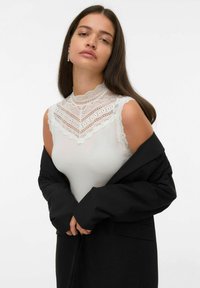 Mujer con cabello largo y oscuro que lleva una blusa sin mangas de encaje blanco y una chaqueta negra sobre los hombros, contra un fondo blanco liso.