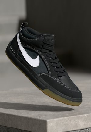 Schwarz-weißer Nike-Sneaker mit Gummisohle, der über einer Betonbrüstung schwebt vor einem verschwommenen dunklen Hintergrund.