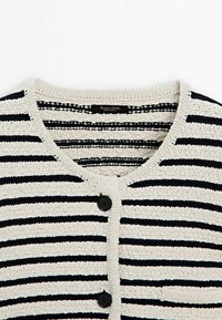 Cardigan beige en tricot avec des rayures horizontales noires, encolure ronde, deux boutons noirs et une petite poche avant.