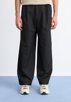Zwarte wijde broek van een glad materiaal, met een voornaad en gecombineerd met beige sneakers. Schoon, minimalistisch ontwerp.