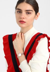 Růžově zlaté náramkové hodinky s kulatým bílým ciferníkem a světle růžovým koženým páskem, které jsou nošeny na ruce. Modelka oblečená v bílém topu s červenými a námořnickými volánky.