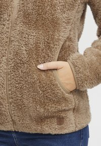 Brown faux fur jacket with a zip front, side pockets, and a textured finish. Features beige trim and a square logo patch at the hem.