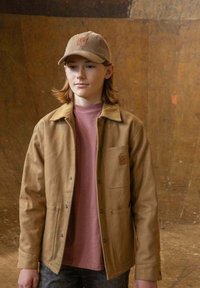 Bruine jas met een kraag, voorzak en drukknoopsluiting, over een lichtroze T-shirt. Draagt een beige corduroy pet.