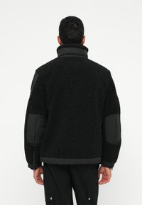 Czarna kurtka polarowa z teksturowaną powierzchnią, wysokim kołnierzem i czarnymi materiałowymi akcentami na rękawach. Posiada zamek błyskawiczny przy mankiecie.