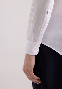 Wit gestreept overhemd met knoopmanchetten. Dunne verticale lijnen, ronde knopen en een klein geborduurd detail op de mouw. Slim fit ontwerp.