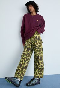 Bordeaux sweatshirt, camouflage-patroon wijde broek in groen en bruin, en zwarte sneakers met groene accenten. Zwarte pet op het hoofd.