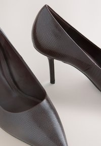 Paire d'escarpins marron foncé à bout pointu avec un motif texturé en peau de serpent sur un fond neutre.