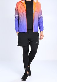 Chaqueta deportiva Nike en degradado de naranja a azul, combinada con pantalones cortos negros y mallas ajustadas. Presenta detalles reflectantes y un diseño elegante.