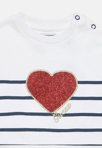 Άσπρο παιδικό μπλουζάκι με οριζόντιες ναυτικές μπλε ρίγες, μια μεγάλη κόκκινη καρδιά με γκλίτερ περιγραμματισμέν η με χρυσό, και το «Guess» κεντημένο με χρυσή γραμματοσειρά.