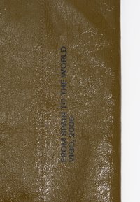 Olivno zelena teksturirana tkanina z bleščečo površino. V mornarsko modri barvi natisnjen tekst pravi "IZ ŠPANIJE V SVET VIGO, 2005," poravnano navpično.