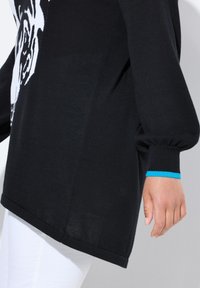 Zwarte gebreide trui met een groot wit grafisch ontwerp, oversized pasvorm en geribde manchetten met een turquoisekleurig accent. Zijdelings een split detail zichtbaar.
