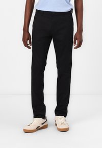 Crne slim-fit hlače izrađene od pamuka, s klasičnim dizajnom od pet džepova i glatkom, mat teksturom. Uparene s bež tenisicama.