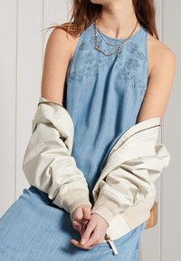 Vestido de mezclilla con bordado floral en el corpiño, llevado con una chaqueta bomber de color crema con puños acanalados y cierre de cremallera.