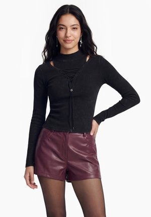 Nuori nainen, jolla on pitkät tummat hiukset, musta pitsinauhoilla kiristettävä pitkähihainen paita, burgundinväriset nahkashortsit ja läpikuultavat mustat sukkahousut, seisoo käsi lanteilla.