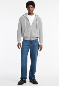 Sudadera gris de canalé con cremallera y capucha, dos bolsillos frontales, combinada con jeans de denim azul rectos y zapatos negros gruesos.