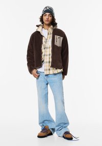 Brązowa polarowa kurtka z beżowym panelem, noszona na koszuli w kratę. Jasnoniebieskie szerokie dżinsy i brązowe sznurowane buty z niebieskimi akcentami.