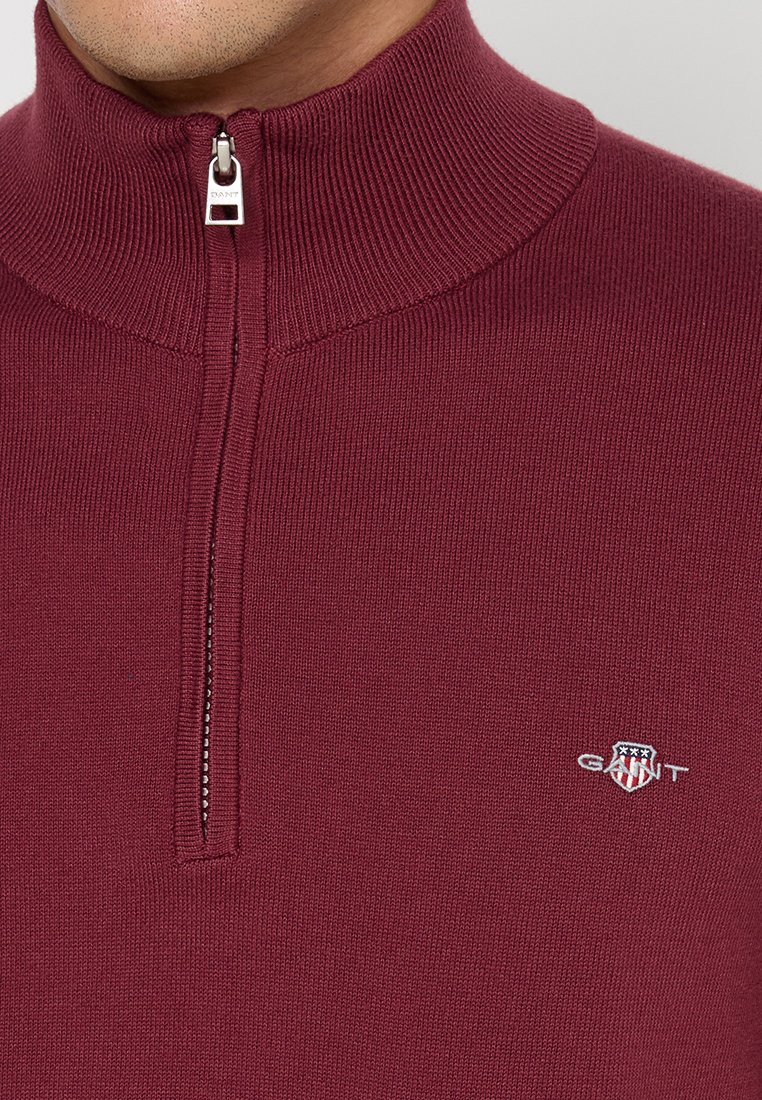 Burgundowy sweter z zamkiem pod szyję, z prążkowanym kołnierzem i małym haftowanym logo na piersi. Ciepły, casualowy styl na chłodniejsze dni.