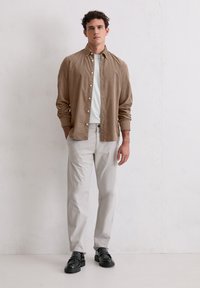 Młody mężczyzna stoi przy białej ścianie, ubrany w beżową koszulę z guzikami na białą koszulkę, jasnoszare spodnie i czarne buty.