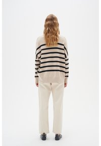 Frau mit welligem blonden Haar, die einen beigen Pullover mit schwarzen Streifen, cremefarbene Hosen und schwarze Schuhe trägt und auf einem weißen Hintergrund von hinten steht.