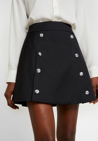 Gonna nera a forma A realizzata in tessuto texturizzato, con bottoni argentati lucidi su un lato, abbinata a una camicia bianca con bottoni.