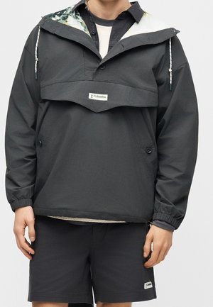 Person trägt eine schwarze Columbia Pulloverjacke mit Frontklappe und schwarze Shorts, stehend vor einem einfarbigen hellen Hintergrund.