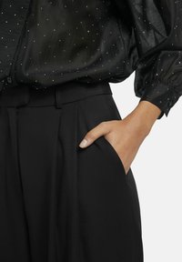 Zwarte, getextureerde blouse met zilveren stippenversiering gecombineerd met hooghangende zwarte broeken; hand in de zak, wat plooien en een gladde stof laat zien.