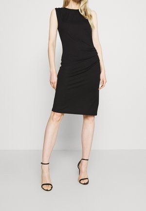 Robe noire sans manches en tissu extensible, avec un design froncé à la taille, associée à des sandales à talons hauts noires à straps.