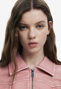 Rosa Kunstlederschnelljacke mit Reißverschluss, spitzem Kragen und detaillierter Näharbeit. Das Model hat lange, wellige braune Haare und natürliche Make-up.