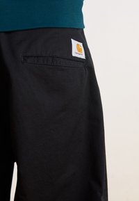 Svarta byxor med en texturerad yta, med en bakficka och en vit etikett med en orange logotyp. Materialet verkar stadigt och hållbart.