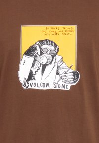 Ruskea t-paita, jossa on graafinen kuva silmälasien kanssa olevasta apinasta keltaisella taustalla, joka pitää puhelinta ja kynää, ja alla on teksti "VOLCOM STONE".