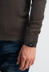 Bruine gebreide trui met een honingraatpatroon, voorzien van een geribbelde zoom en manchetten. Het label op de mouw zegt "Butcher of Blue." G gedragen met denim.