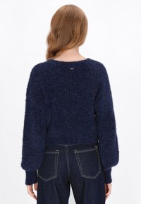 Granatowy, puszysty sweter z dzianiny z szerokim okrągłym dekoltem i balonowymi rękawami. Posiada prążkowane mankiety i krótkie wykończenie u dołu. Minimalne oznakowanie z tyłu.