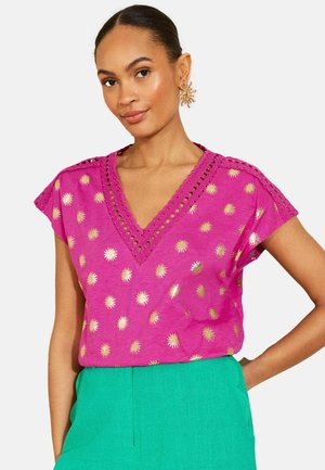 Žena nosiaca magentové tričko s výstrihom do V so zlatými slnečnými vzormi a zelené nohavice, doplnené zlatými náušnicami v tvare slnka.