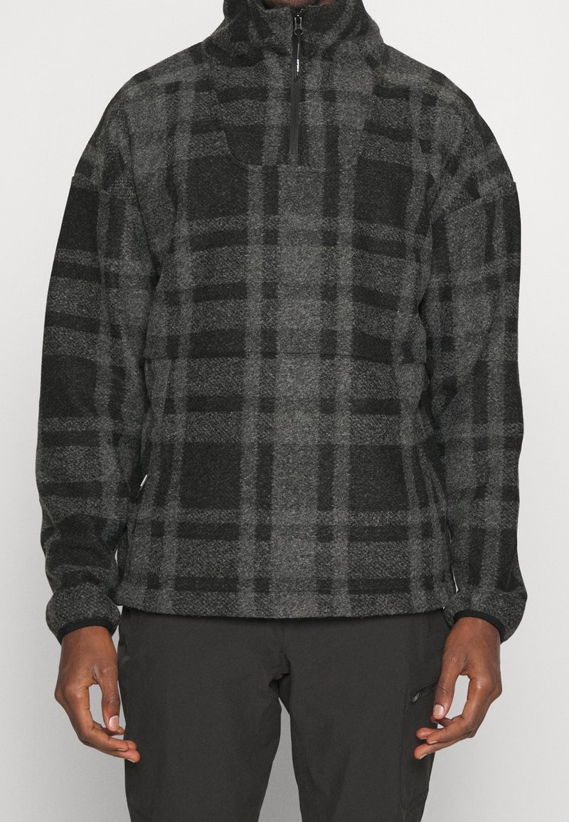 Homme portant un pullover polaire à carreaux gris foncé et noir avec une fermeture éclair quart de longueur et un pantalon noir, debout, les bras détendus le long du corps.