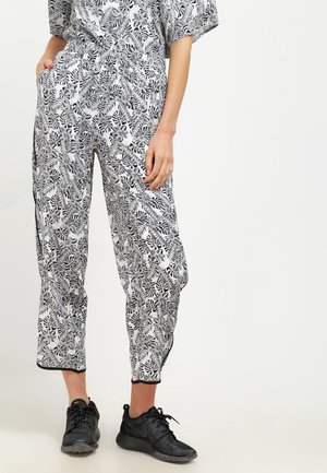 Kvinde iført sort-hvide bukser med bladmønster og sorte sneakers, stående med den ene hånd i lommen mod en ensfarvet baggrund.