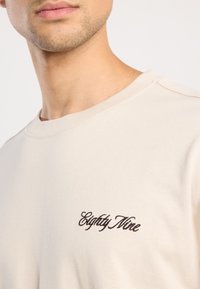 Beige puuvillane T-särk, millel on ümar kaelus ja pruuni tikandiga "Eighty Nine" logo vasakul küljel. Sile tekstuur, casual disain.