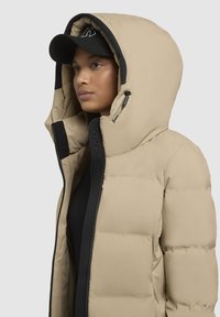 Beige pufferjack met een zwarte rits en accenten. Heeft een grote capuchon en een gestructureerde stof. Onder de jas is een zwart top zichtbaar.