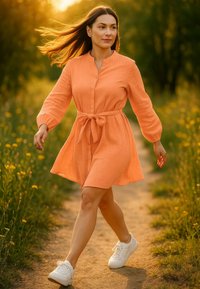 Abito arancione con bottoni e cintura, maniche lunghe e tessuto texturizzato; indossato con sneakers bianche, posato su un sentiero erboso.