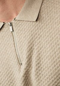 Beige-vetoketjullinen poolopaita, joka on valmistettu teksturoidusta kankaasta ja jossa on timanttikuvio. Paidassa on kaulus ja hopeanvärinen vetoketjusomiste.