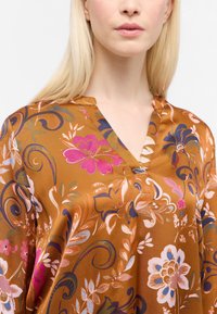 Brun bluse med mønster, der har blomster i pink, lilla og hvid. Blødt, glat stof med V-udskæring og korte knapdetaljer.