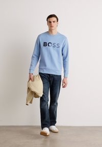 Light blue sweatshirt with "BOSS" logo, dark denim jeans, white sneakers, and a beige jacket in hand. Simple, casual outfit design.