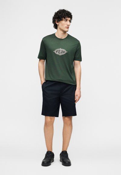 Άνδρας με σγουρά μαλλιά που φοράει πράσινο κοντομάνικο πουκάμισο με λογότυπο, μαύρο σορτς και μαύρα αθλητικά παπούτσια, στέκεται με τα χέρια στις τσέπες.