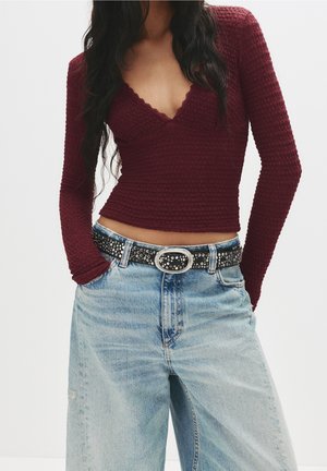 Femme portant un haut bordeaux texturé à manches longues avec un profond décolleté en V, un jean taille haute bleu clair et une ceinture noire cloutée.