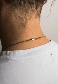 Gul snoet kædedesign med en lås, hviler på bar hud ved nakken. Hvid skjortekrave synlig i baggrunden.