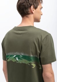 Olijfgroene katoenen t-shirt met een ronde hals en korte mouwen. Bevat een landschapsgrafiek in tinten groen en lichtblauw over de rug.
