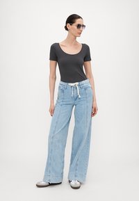 Grå scoop-neck topp kombinert med lyseblå vide jeans med snøring. Modell med solbriller og casual sko, stående oppreist.