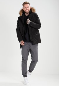 Svart parkajakke med fôr i hetta, glidelås og knappelukking, to framlommer, båret over en svart genser og grå bukser, med hvite joggesko.