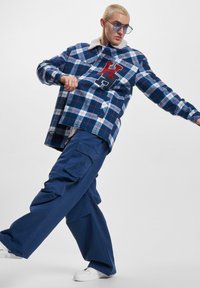 Ternet flannel skjorte i navy og hvid, med rød bogstavs-patch. Kombineret med cargo bukser, fremhævet af hvide sneakers.
