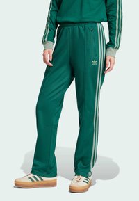 Gröna sweatpants med avslappnad passform, med tre vertikala ljusare gröna ränder på varje sida och en blixtlåsficka på vänster ben.