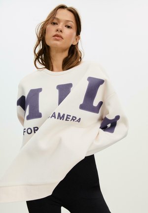 Felpa color crema chiaro con lettering viola bold. Presenta una vestibilità ampia, maniche lunghe e un collo rotondo con bordo a costine.